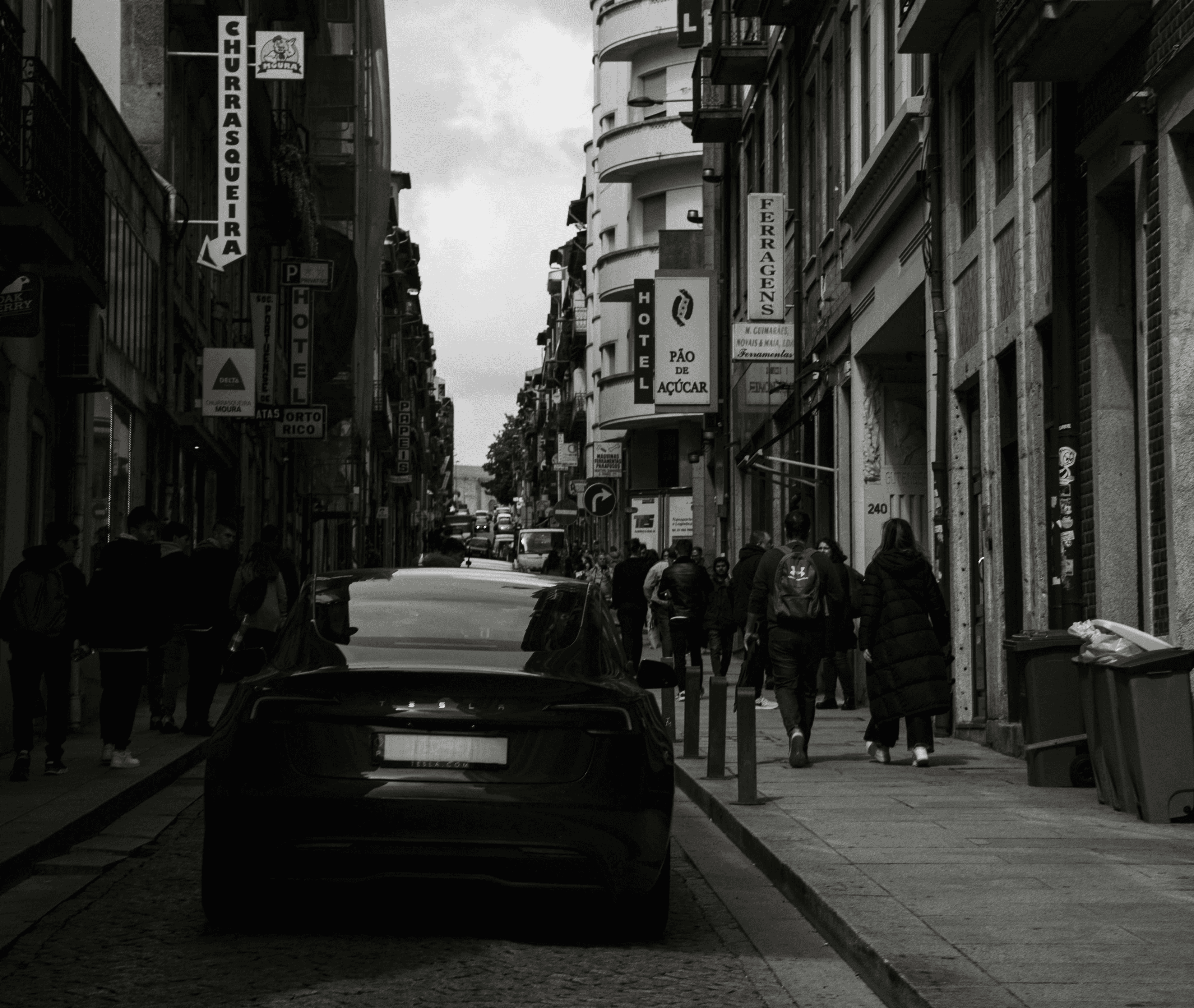 Carro elétrico visto de trás numa rua urbana de Portugal, em preto e branco, com peões ao fundo